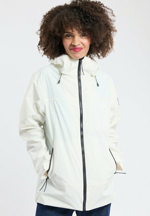 Femme aux cheveux bouclés portant une veste blanche zippée, les mains dans les poches, debout devant un fond clair uni.