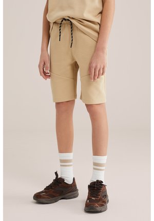 Laps, kes kannab beeže põlvpikkuseid lühikesi pükse musta pingutuspaelaga, valgeid sokke beežide triipudega ning pruunide massiivsete tossudega, seisab neutraalsel taustal.