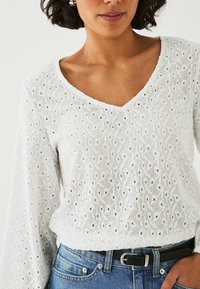 Blusa bianca ricamata con scollo a V, maniche lunghe e intricati motivi floreali con fori a occhio. Abbinata a jeans in denim blu.