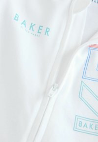Balta zip-up gobtuvė su "BAKER" logotipu šviesiai mėlynos spalvos ir spalvingais geometriniais raštais. Pagaminta iš medvilnės mišinio, turinčio lygią tekstūrą.