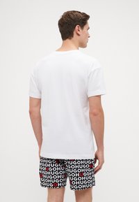 T-shirt blanche à manches courtes associée à un short noir présentant un texte rouge et blanc qui dit "HUGO" dans un motif répétitif.