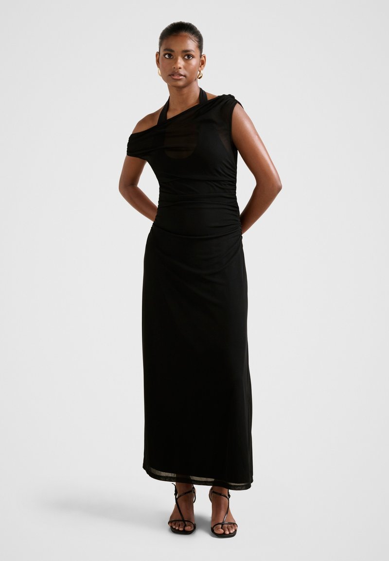 Kobieta ubrana w długą czarną suknię na jedno ramię z asymetrycznym dekoltem i czarne sandały na obcasie, stojąca z rękami za plecami.