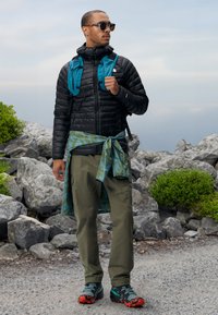Zwarte pufferjack, turquoise rugzak, groene shirt met patroon om de taille gebonden, olijfkleurige cargobroek en meerkleurige wandelschoenen.