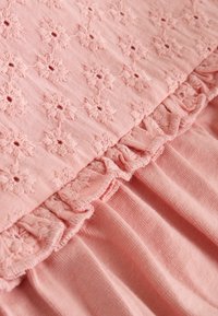 Rózsaszínes textília virágos hímzéssel és fodros szegéllyel. Puha tapintású, réteges kialakítás, varrott és sima részek keverékével.