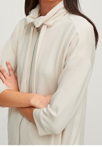 Lyse beige jakke med høj krave, korte ærmer og lynlås foran. Glat stof med en blød tekstur.