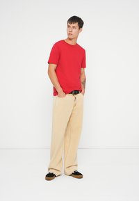 T-shirt de algodão vermelho combinado com calças largas bege. Footwear apresenta detalhes em preto e branco, com uma sola texturizada. Modelo está em um fundo neutro.