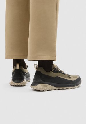 Person, die beige weit geschnittene Hosen und schwarze sowie olivgrüne Wanderschuhe mit groben Sohlen trägt, steht auf einem schlichten hellen Hintergrund.