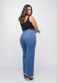 Vrouw met lang golvend haar draagt een zwarte mouwloze top, blauwe high-waisted jeans en zwarte hoge hakken, poserend tegen een witte achtergrond.