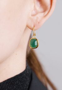 Cercei inelari de culoare aurie cu o piatră verde fațetată. Piatra are formă ovală, fixată într-un bezel neted din aur. Urechea texturată și îmbrăcămintea sunt vizibile.
