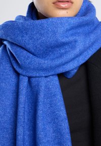 Blue wool scarf with a soft texture, featuring a woven pattern. Draped loosely around the neck, partially covering a black garment.