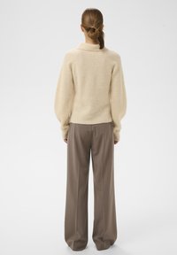 Sieviete ar savelktaem matiem, ģērbusies bēšā adītā džemperī un brīvās brūnās biksēs, stāvot ar muguru pret vienkāršu balto fonu.