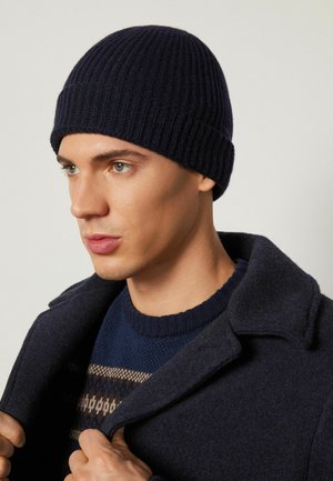 Beanie a coste blu navy, aderente con risvolto. Indossato con un cappotto scuro e un maglione con motivo, caratterizzato da tonalità e texture terrose.