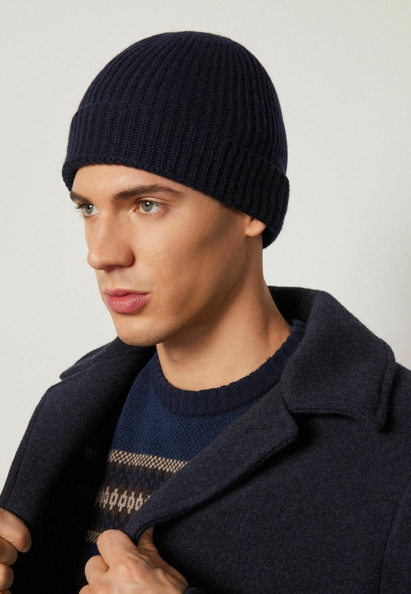 Marinblå ribbad stickad mössa, åtsittande med en vikbar kant. Bärs med en mörk rock och mönstrad tröja i jordnära toner och texturer.