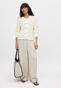 Cardigan en crème texturé avec un col rond et des boutons, associé à un pantalon beige ample. Porte un sac fourre-tout structuré beige et noir.