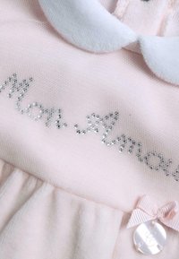 Lyserød kjole med hvid krave, prydet med glitrende perler, der staver "Mon Amour", en lille sløjfe og en dekorativ knapdetalje. Blød tekstur.