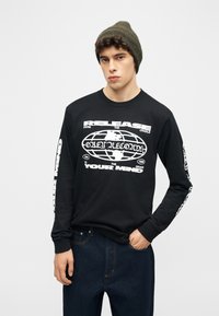 Νέος άνδρας που φοράει πράσινο σκουφάκι και μαύρο μακρυμάνικο πουκάμισο με λευκό γραφικό παγκόσμιου χάρτη και κείμενο, στέκεται με το ένα χέρι στην τσέπη.