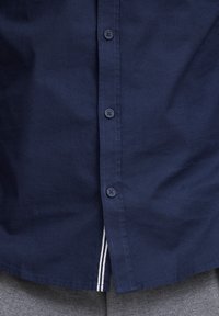 Camisa de botones azul marino, hecha de algodón, con cinco botones y una franja blanca en el dobladillo inferior. Textura suave.