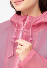 Giacca da pioggia traslucida rosa con cappuccio, caratterizzata da una cerniera frontale, cordini e polsini con bottone a pressione. Texture liscia e design leggero.
