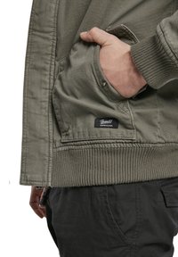 Olivgrüne Jacke mit gerippten Bündchen, ausgestattet mit einer Seitentasche und einem schwarzen Etikett. Der Stoff ist glatt und hat einen subtilen Glanz.