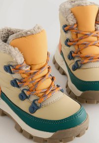 Bottes isolées beige et orange avec une bordure en fourrure texturée, des lacets bleus et orange, et des semelles robustes vertes et blanches. Œillets renforcés visibles.