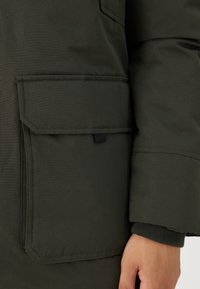 Green, insulated jacket featuring a large flap pocket with a black snap closure, ribbed cuffs, and stitched seams, made from durable fabric.