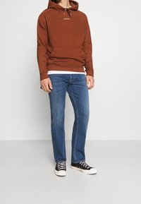 Felpa cappuccio marrone con tasca frontale e logo bianco; abbinata a jeans blu e sneaker nere con suole bianche.