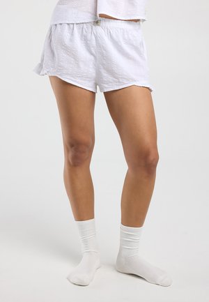Jambes portant un short blanc texturé et un haut assorti, avec des chaussettes blanches côtelées, debout devant un fond clair uni.