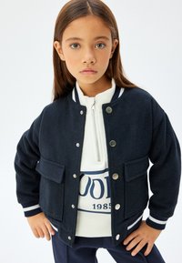 Giacca bomber in lana blu navy con due tasche, bottoni a pressione e polsini a righe. Indossata sopra una maglietta bianca con zip caratterizzata da una grafica blu.