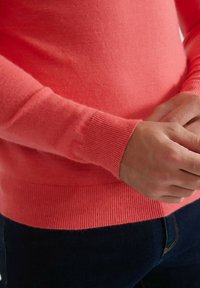Pull couleur corail en tissu doux, avec des poignets et un ourlet côtelés. Gros plan montrant la texture lisse et le design ajusté.