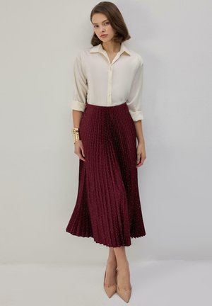 Femme debout portant une chemise crème boutonnée, une jupe midi plissée bordeaux à petits pois, des talons pointus beiges et des bracelets dorés au poignet gauche.