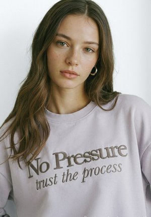 Giovane donna con lunghi capelli castani, che indossa orecchini a cerchio e una felpa lavanda con la scritta "No Pressure trust the process."