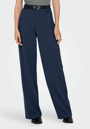 Femme portant un pantalon large bleu marine associé à un haut gris sans manches, une ceinture noire et des chaussures noires à bout pointu sur un fond uni.