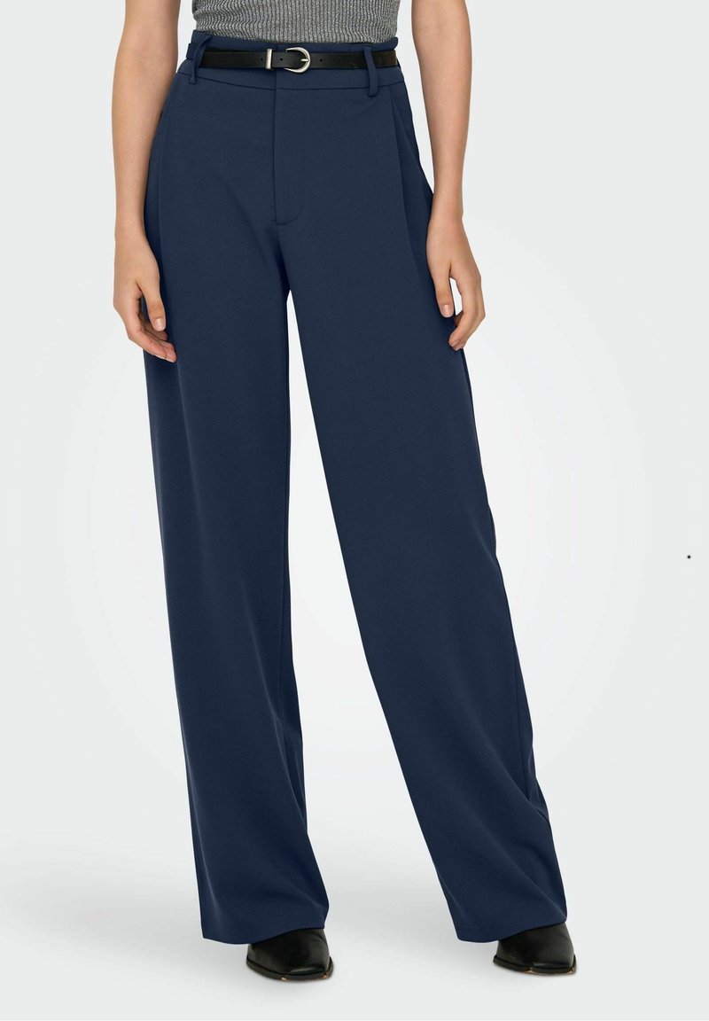 Femme portant un pantalon large bleu marine associé à un haut gris sans manches, une ceinture noire et des chaussures noires à bout pointu sur un fond uni.