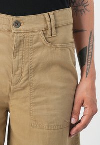 Pantaloni di cotone beige con una tasca frontale, dettagli di cucitura visibili e chiusura con bottone metallico in vita.