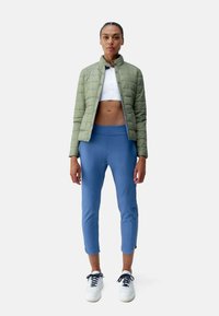 Lichtgroene gewatteerde jas, witte crop top en blauwe aansluitende broek met enkel lengte, gecombineerd met witte sneakers en donkere veters.