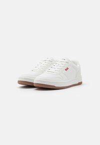 Weiße Sneakers aus strukturiertem Leder, runde Zehenpartie, Gummisohle und leichtem Design. Mit subtil rotem Logo an der Seite und weißen Schnürsenkeln.
