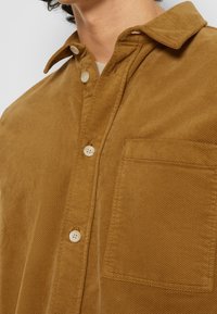 Gros plan d'une personne portant une chemise en velours côtelé marron avec des boutons beige et une poche poitrine, montrant une partie du col et du cou.