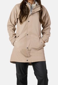 Imperméable beige clair avec capuche, fermeture éclair à pression à l'avant et deux poches latérales. Présente une texture lisse et des cordons réglables.