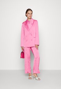 Blazer de satin rosa com botões em forma de coração, combinado com calças correspondentes. O conjunto inclui uma camisa com colarinho e sandálias brancas de salto alto.