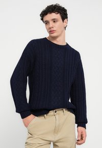 Πλεκτό μπλε πουλόβερ σε σκούρο ναυτικό μπλε με υφασμένο σχεδιασμό καλωδίου, με ριγωτές μανσέτες και τελείωμα, εμφανίζεται με ανοιχτόχρωμο παντελόνι.