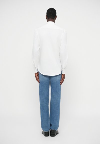 Λευκό πουκάμισο μακρύ μανίκι με μανσέτες κουμπιών, μπλε τζιν παντελόνι και μαύρα παπούτσια, φαίνεται από πίσω σε έναν απλό λευκό φόντο.