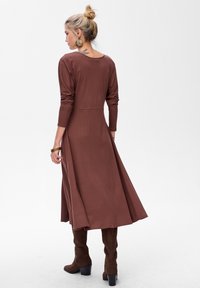 Robe longue marron à manches longues avec un corsage ajusté et une jupe évasée, confectionnée dans un tissu doux. Associée à des bottes hautes en suède marron.
