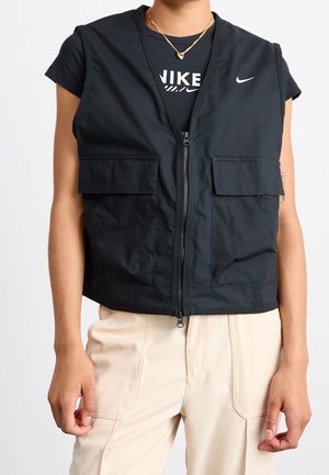 Personne portant un gilet zippé noir Nike par-dessus un t-shirt noir Nike, associé à un pantalon beige sur un fond uni.
