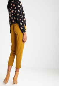 Svart blommig topp med lång ärm, ihop med senapsgula tapered byxor och bruna ankelboots, vilket framhäver en livlig färgkontrast.