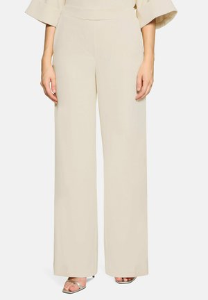 Vrouw draagt crèmekleurige broek met hoge taille en wijde pijpen, gecombineerd met zilveren open-toe hakken, staand tegen een effen achtergrond.