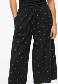 Melni plaši bikses, kas izgatavotas no mīksta auduma, ar dimanta rakstu un sānu kabatām, kā arī ar elastīgu jostu komfortam.