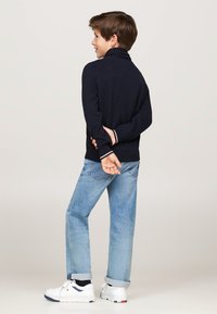 Maglione navy con colletto alto, maniche lunghe e dettagli bianchi ai polsini, abbinato a pantaloni in denim azzurro chiaro e sneakers bianche.