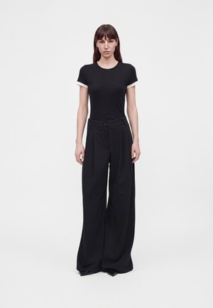 Frau mit langen braunen Haaren, die ein schwarzes, eng anliegendes T-Shirt mit weißem Besatz und weite schwarze Hosen trägt, steht vor einem schlichten weißen Hintergrund.