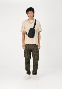 Zwarte crossbodytas gedragen op een beige t-shirt, gecombineerd met groene camouflage cargobroek en witte sneakers. De tas heeft een rits en een verstelbare schouderband.