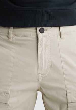 Beige broek met knoopsluiting, riemlussen en zichtbare stiksels, gedragen onder een zwarte geribbelde trui.